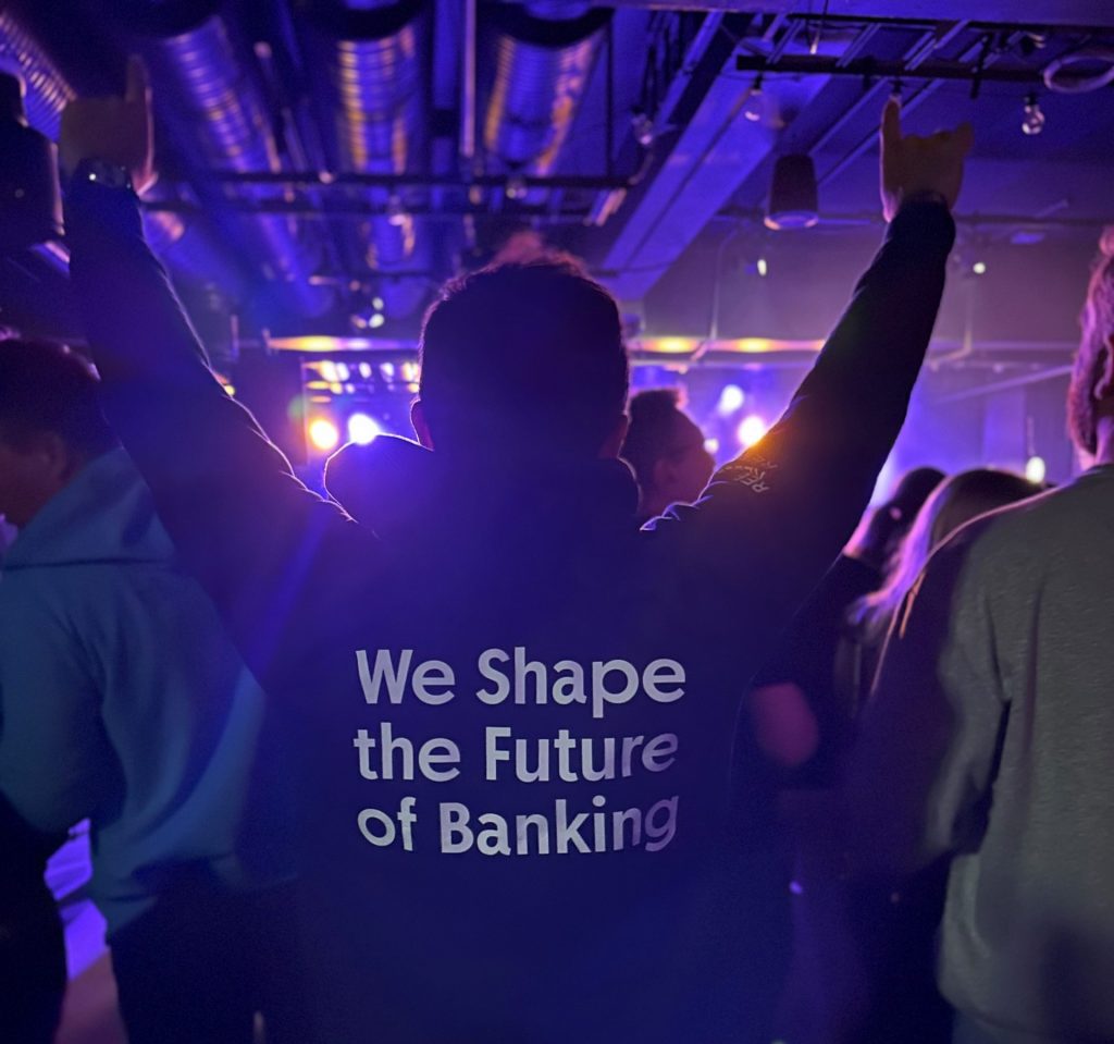 Man standing with his hands in the air. He's wearing a hoodie with 'We Shape the Future of banking' written on it.